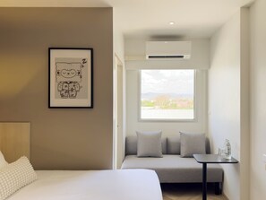 Una habitación de hotel moderna con cama, sofá, una mesita con vasos y botellas de agua, un cuadro en la pared y un ventanal con vistas a la ciudad.