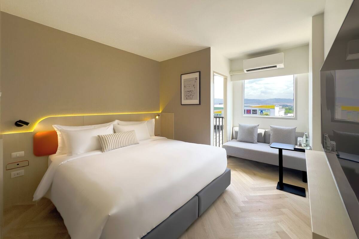 A modern hotel room with a large bed, a sofa, a dining table, and a view of the city.