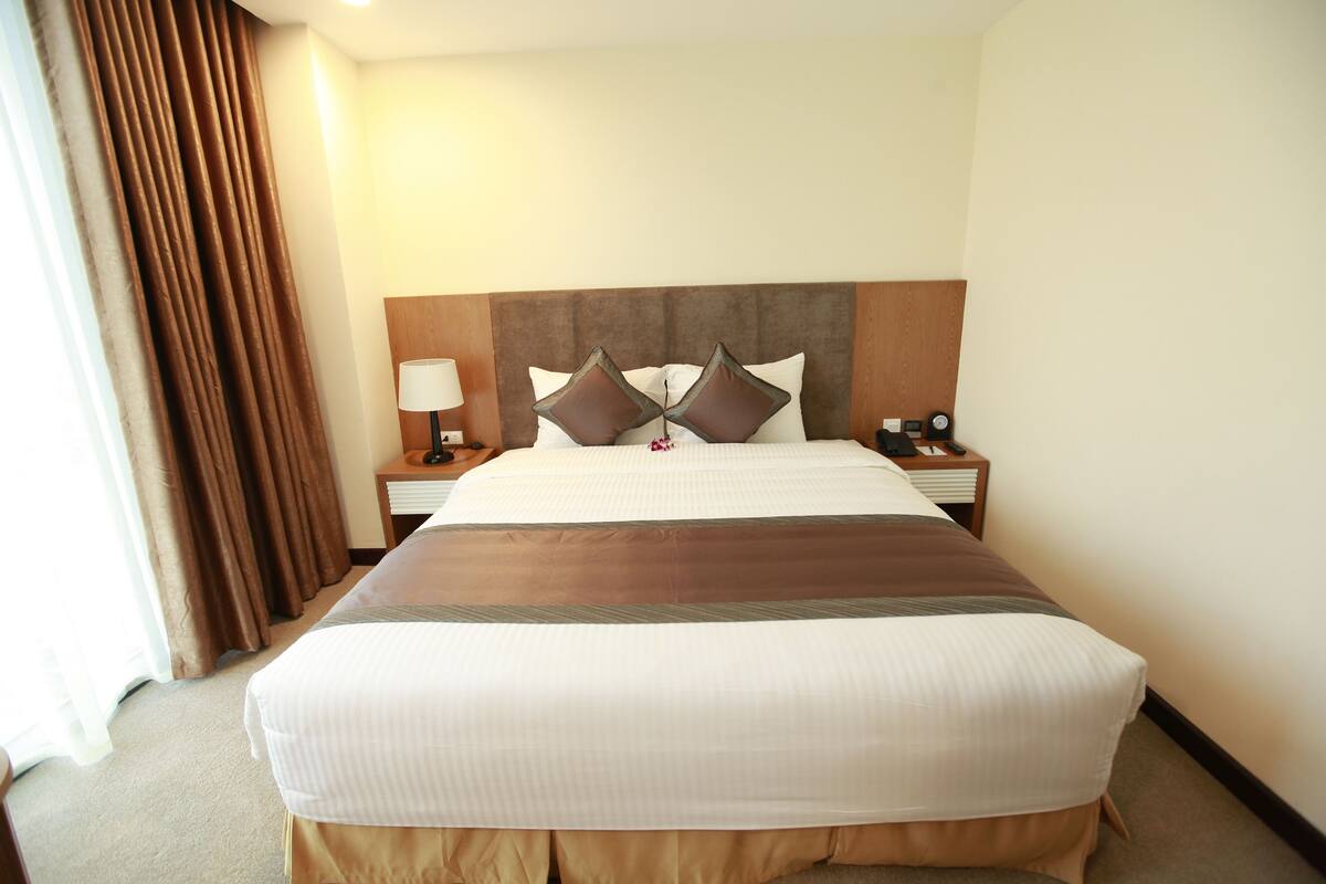 Une chambre d’hôtel avec un grand lit, une tête de lit en bois, une table de chevet avec une lampe et une fenêtre avec des rideaux.