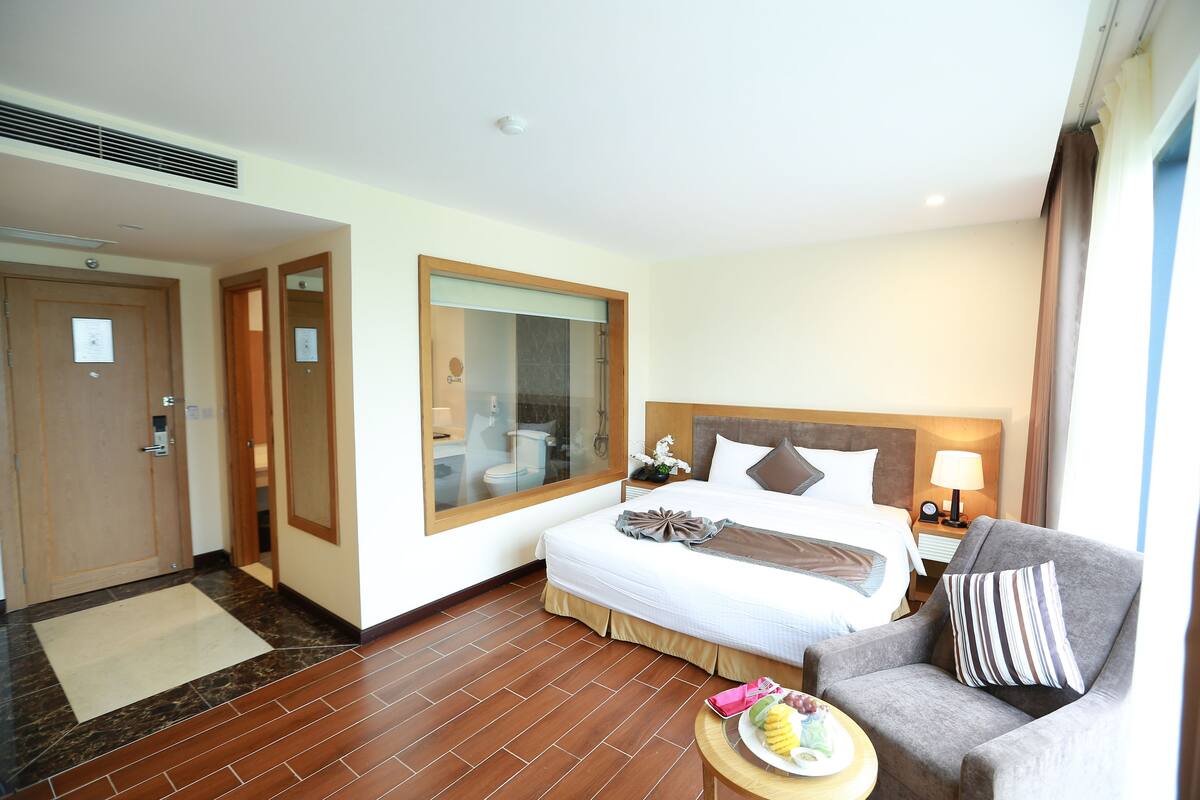 Une chambre d’hôtel avec un lit, une chaise, une petite table avec des fruits, et une salle de bain visible à travers une porte en verre.