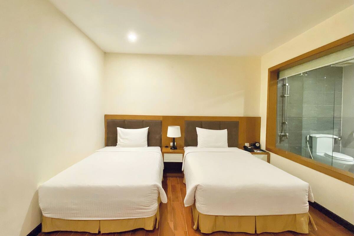 Une chambre d’hôtel avec deux lits simples, un plancher en bois, une table de chevet avec une lampe et une fenêtre donnant sur une douche.