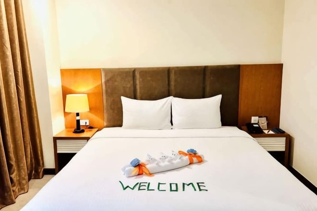 A hotel room with a bed, a nightstand with a lamp, and a welcome mat.