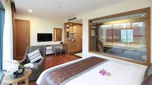 Une chambre d’hôtel moderne comprenant un lit, un coin salon avec un fauteuil et une petite table, une télévision et une grande fenêtre donnant sur l’extérieur.