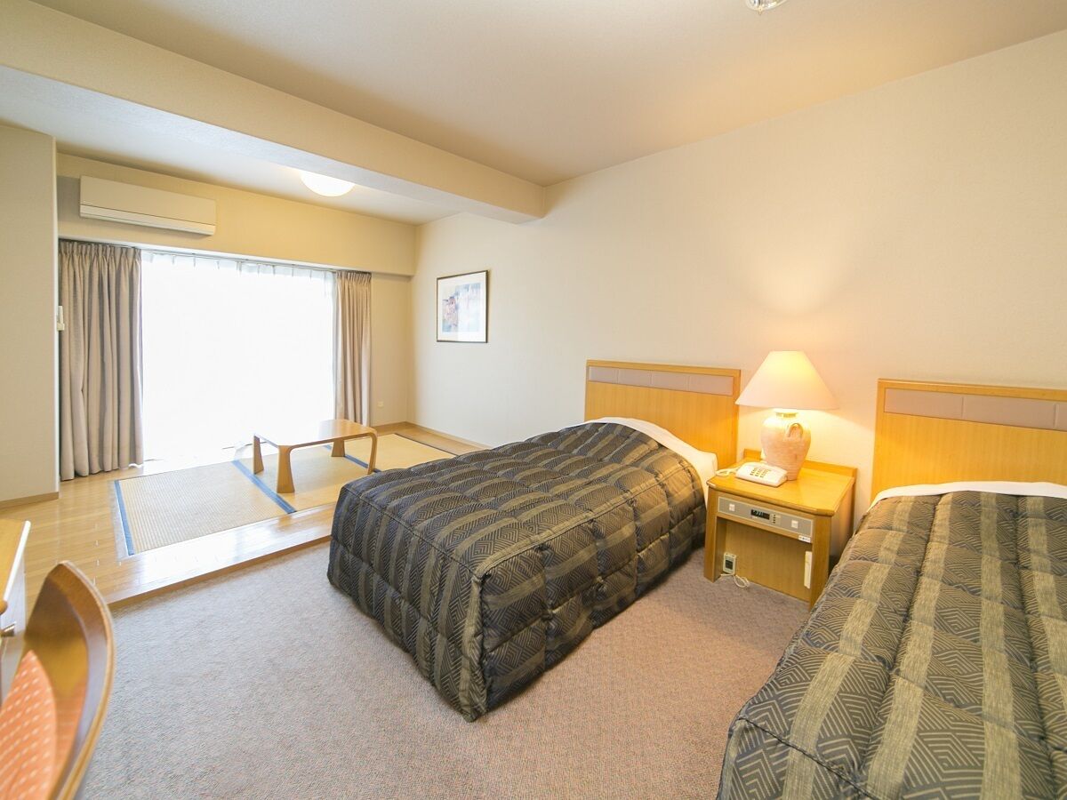 A hotel room with a bed, bedside table with lamp, and a window with curtains.