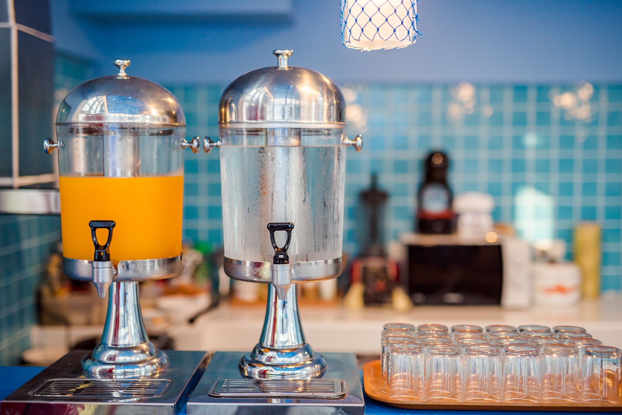 Two stainless steel dispensers with transparent yellow liquid, a tray of clear glasses, and a blue tiled backsplash in the background.