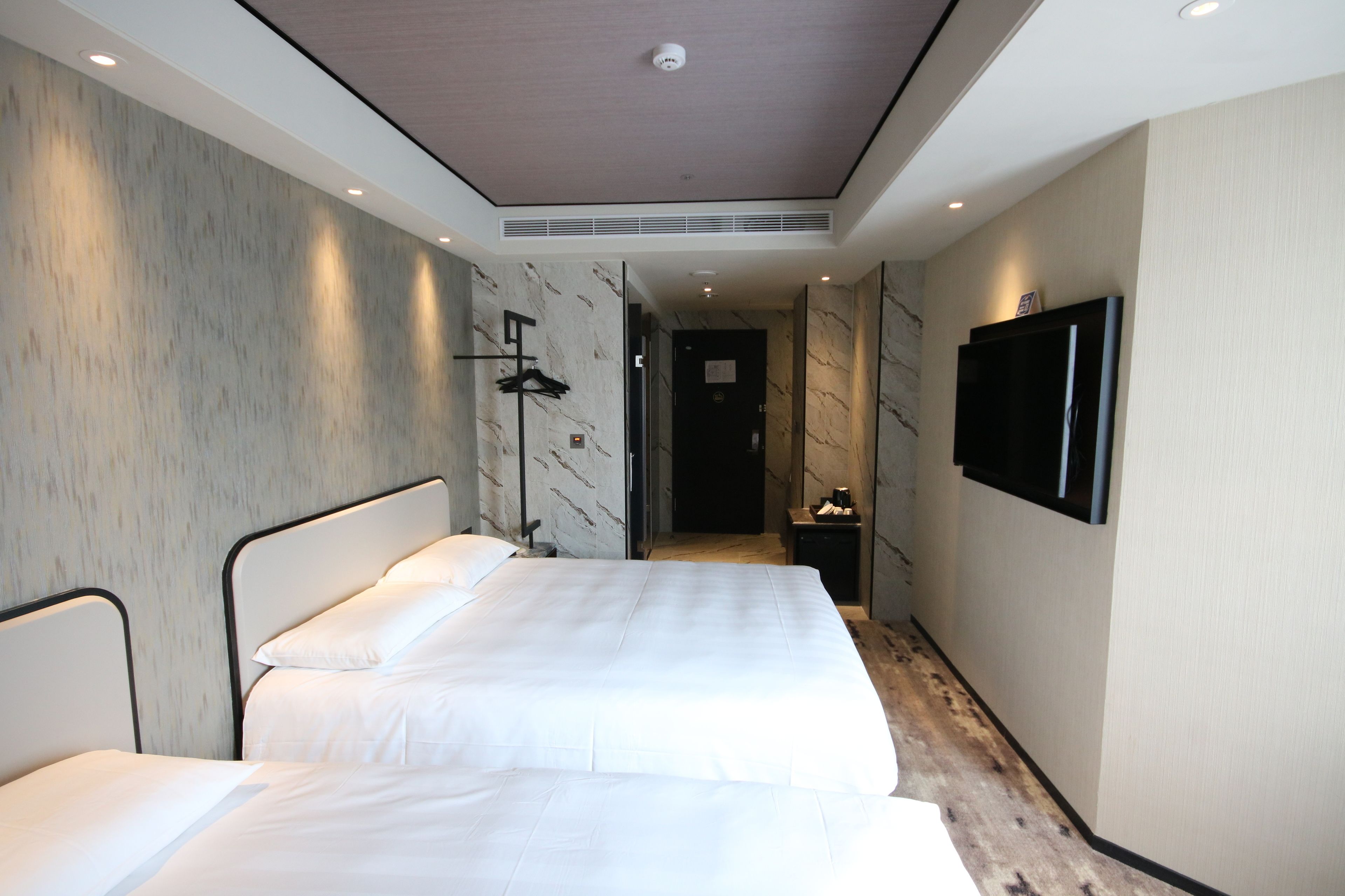 A hotel room with two beds, a flat-screen TV mounted on the wall, and a hallway leading to a door.
