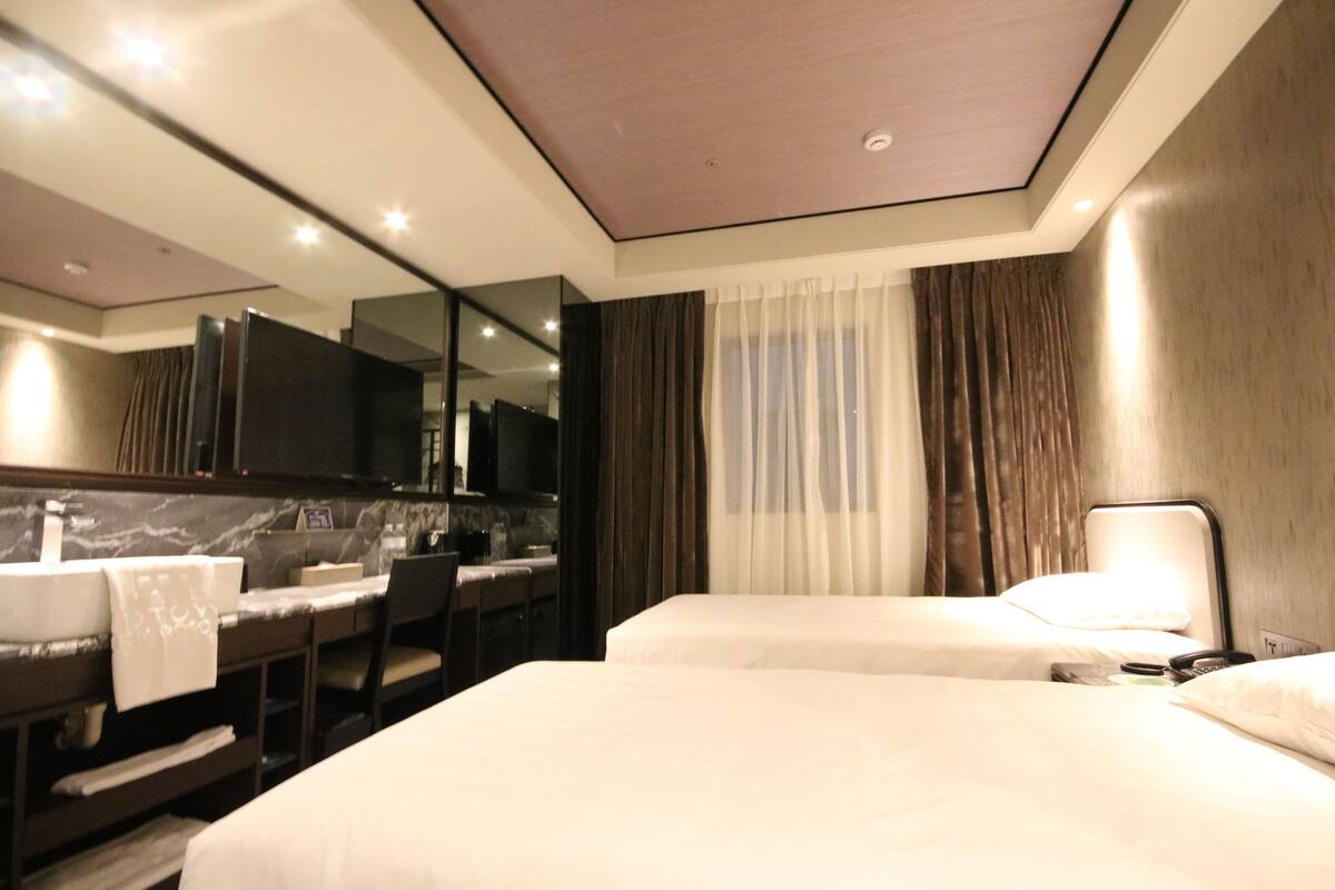A hotel room with two beds, a desk with a chair, a sink, and a large mirror.