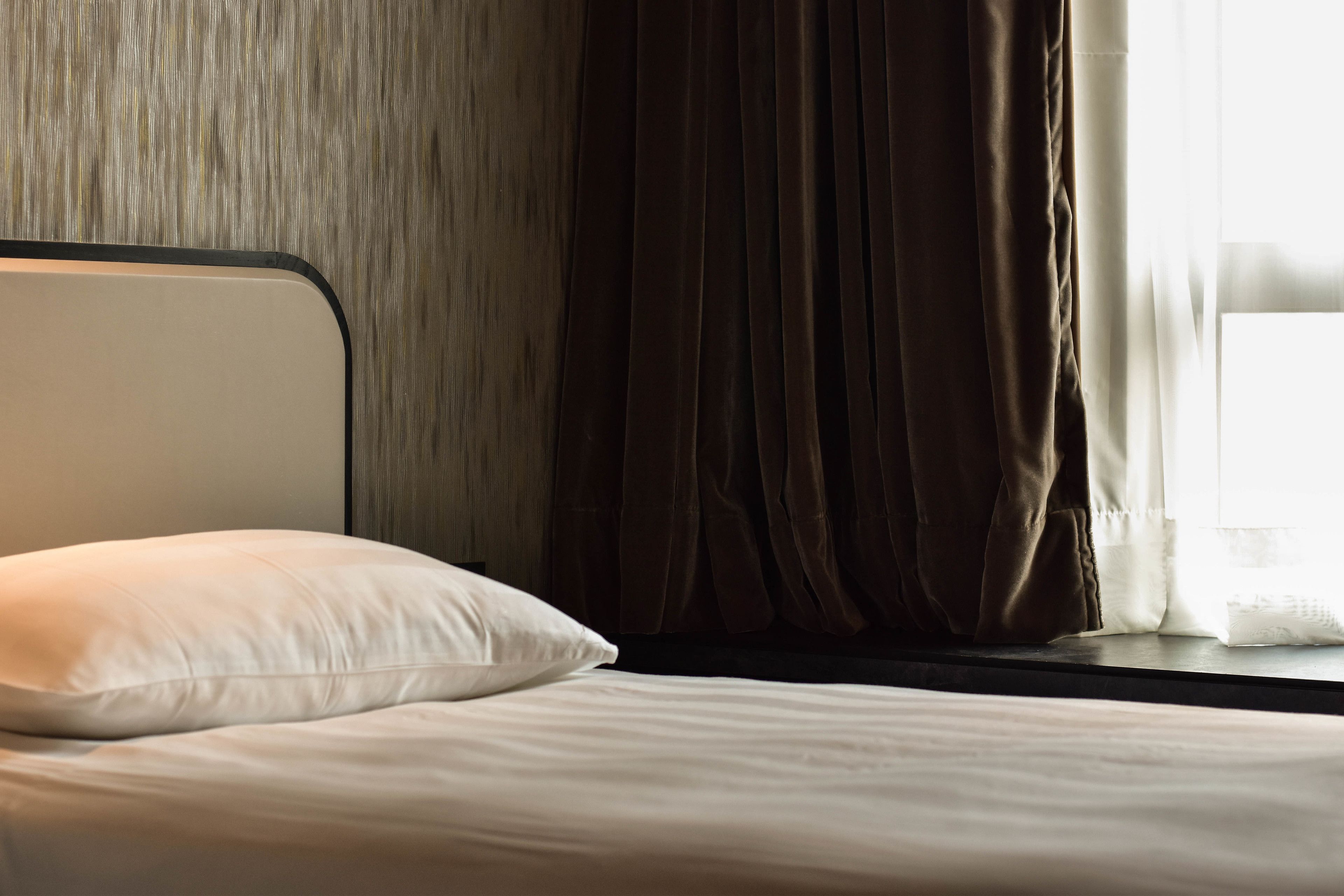 Una cama con un almohadón blanco y un cabecero, una ventana con cortinas y una pared de madera.