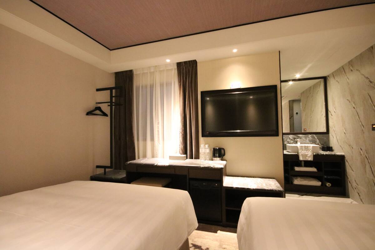 A hotel room with two beds, a desk, a flat-screen TV, a mirror, and a bathroom with a marble wall.