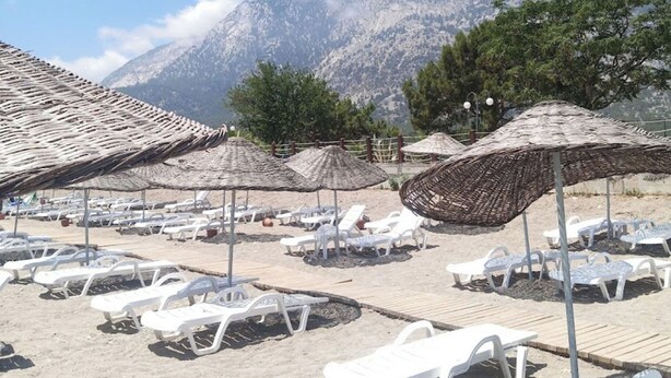 A beach with white lounge chairs and straw umbrellas, with mountains in the background. - Beldibi Beach Hotel (Kemer)