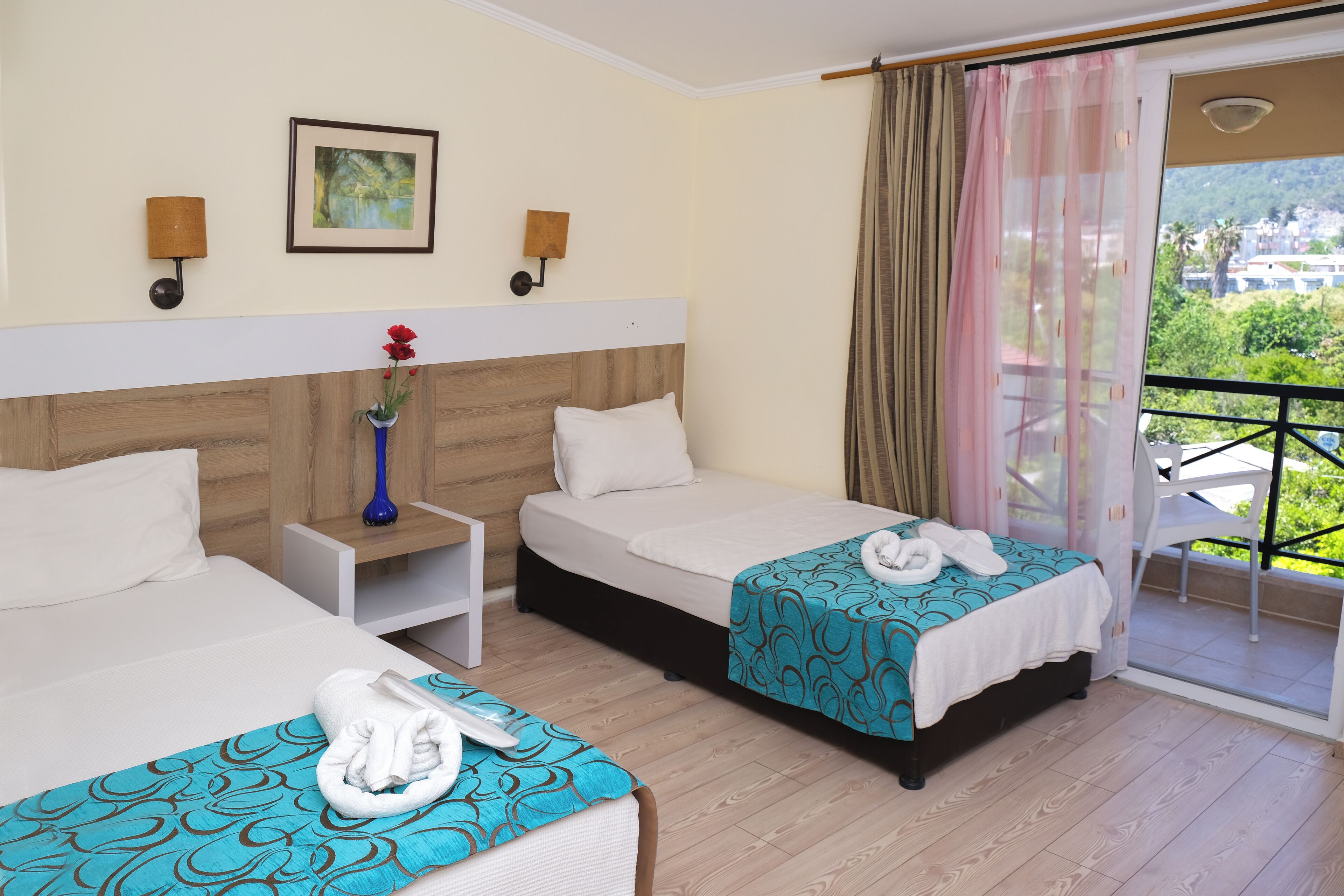 Une chambre d’hôtel avec deux lits, une table de chevet, un vase avec une fleur rouge et un balcon offrant une vue sur la verdure et les bâtiments.