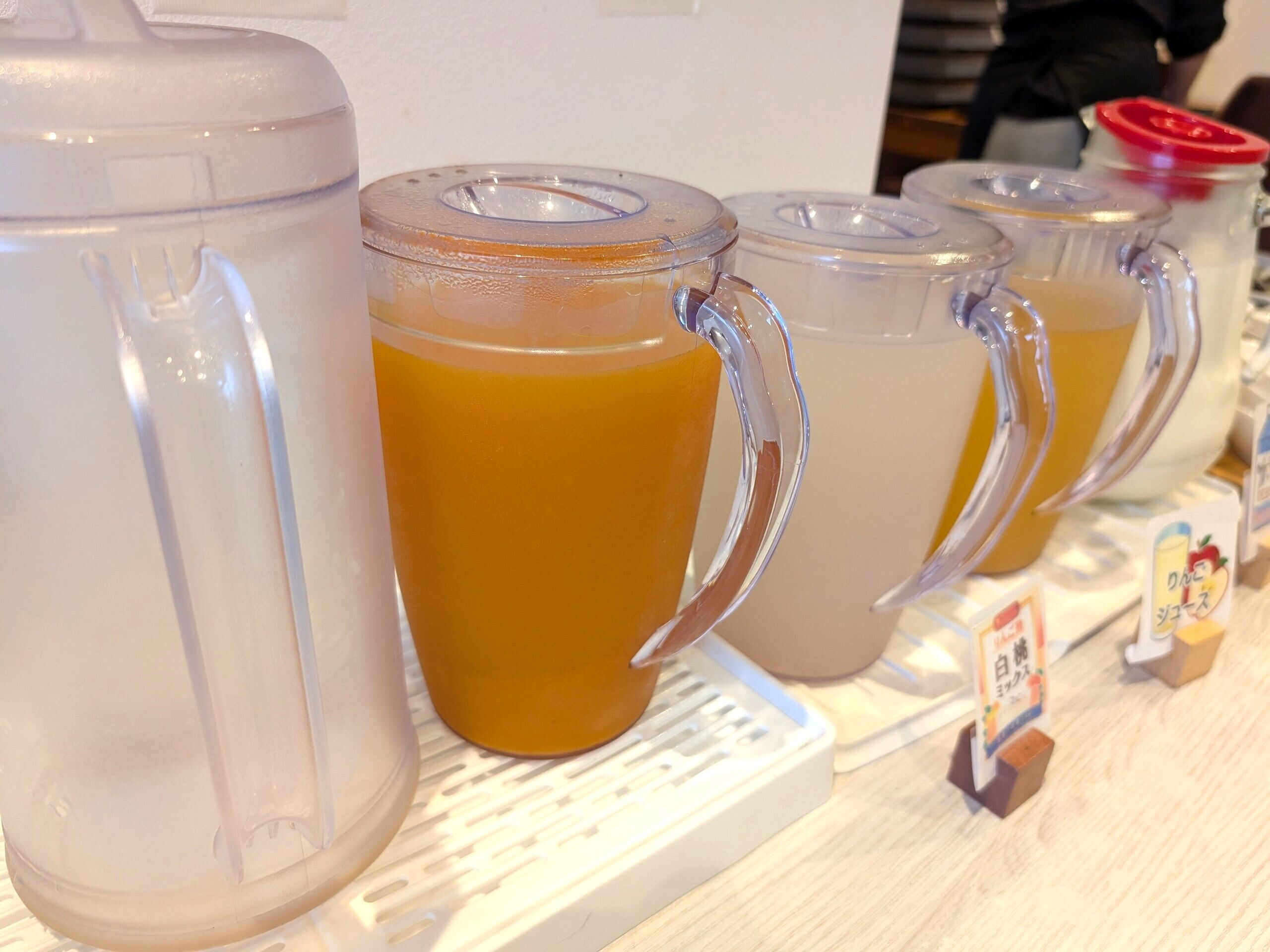 Une rangée de verres transparents à anse, remplis d’un liquide jaunâtre, vraisemblablement une boisson, sur une étagère blanche.
