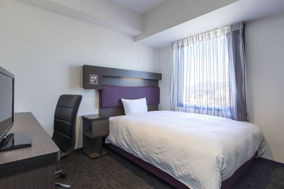 A hotel room with a bed, a desk with a computer, a chair, and a TV.