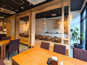 Un interior de restaurante moderno con mesas y sillas de madera, iluminación colgante y un ventanal con una planta.