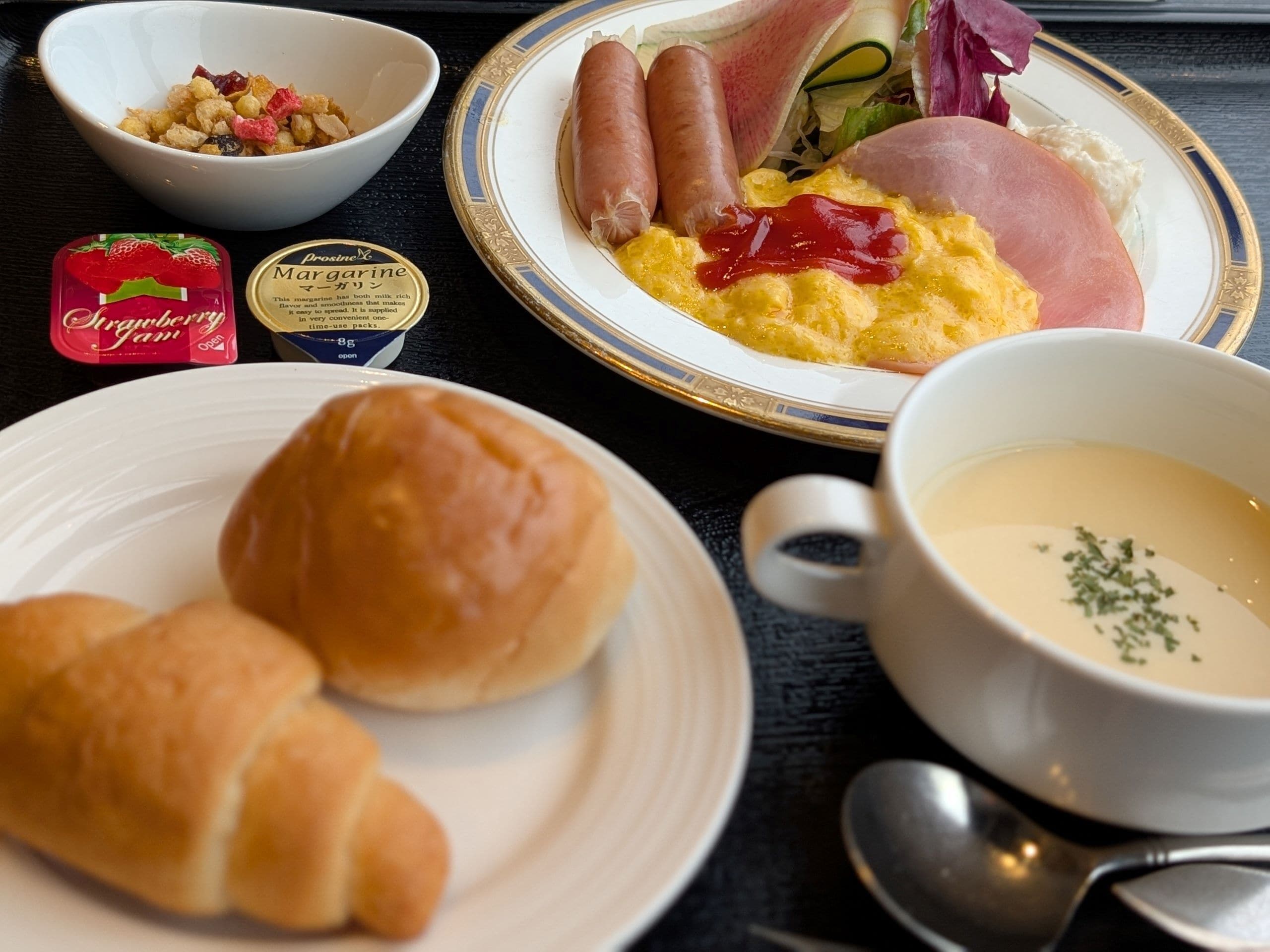A meal with sausages, ham, scrambled eggs, and a bowl of soup.