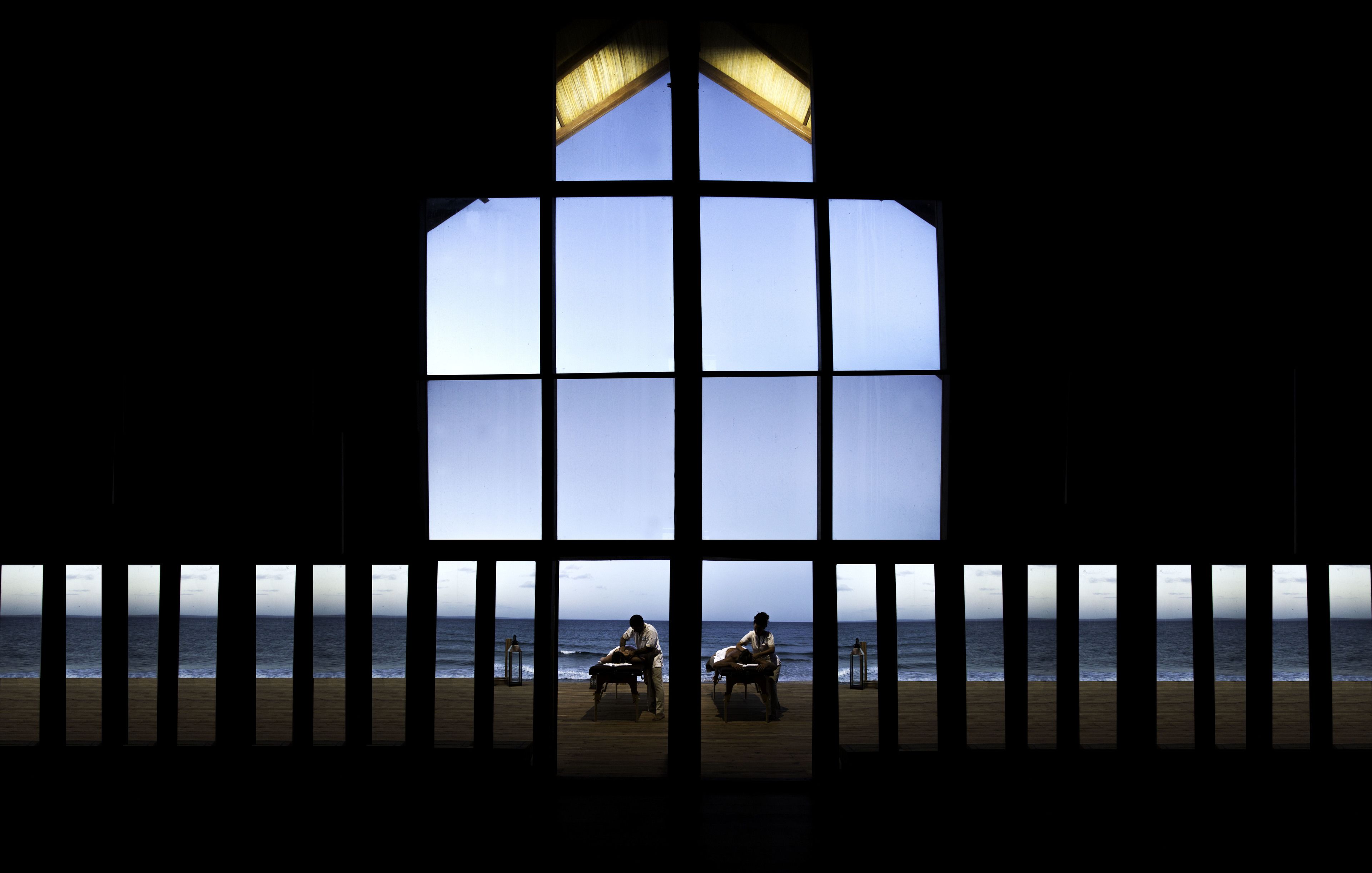 A room with a large window overlooking a beach, featuring a wooden floor and a view of the sea.