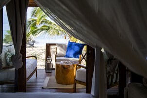 Una habitación frente a la playa con una cama, una silla de madera con un cojín azul, una mesa redonda de madera y vistas a palmeras y la playa.