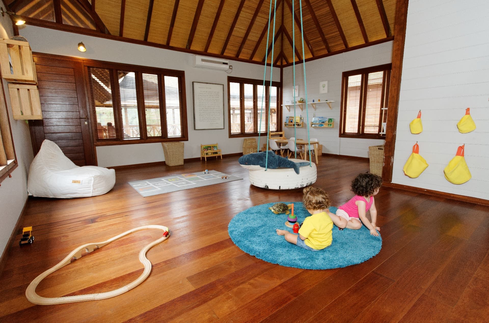 Una habitación amplia con suelo de madera, alfombra azul, una silla colgante y un área de juegos con juguetes.