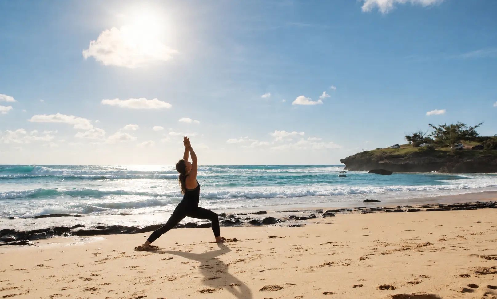A person performing yoga on a sandy beach with the ocean and a rocky outcrop in the background.