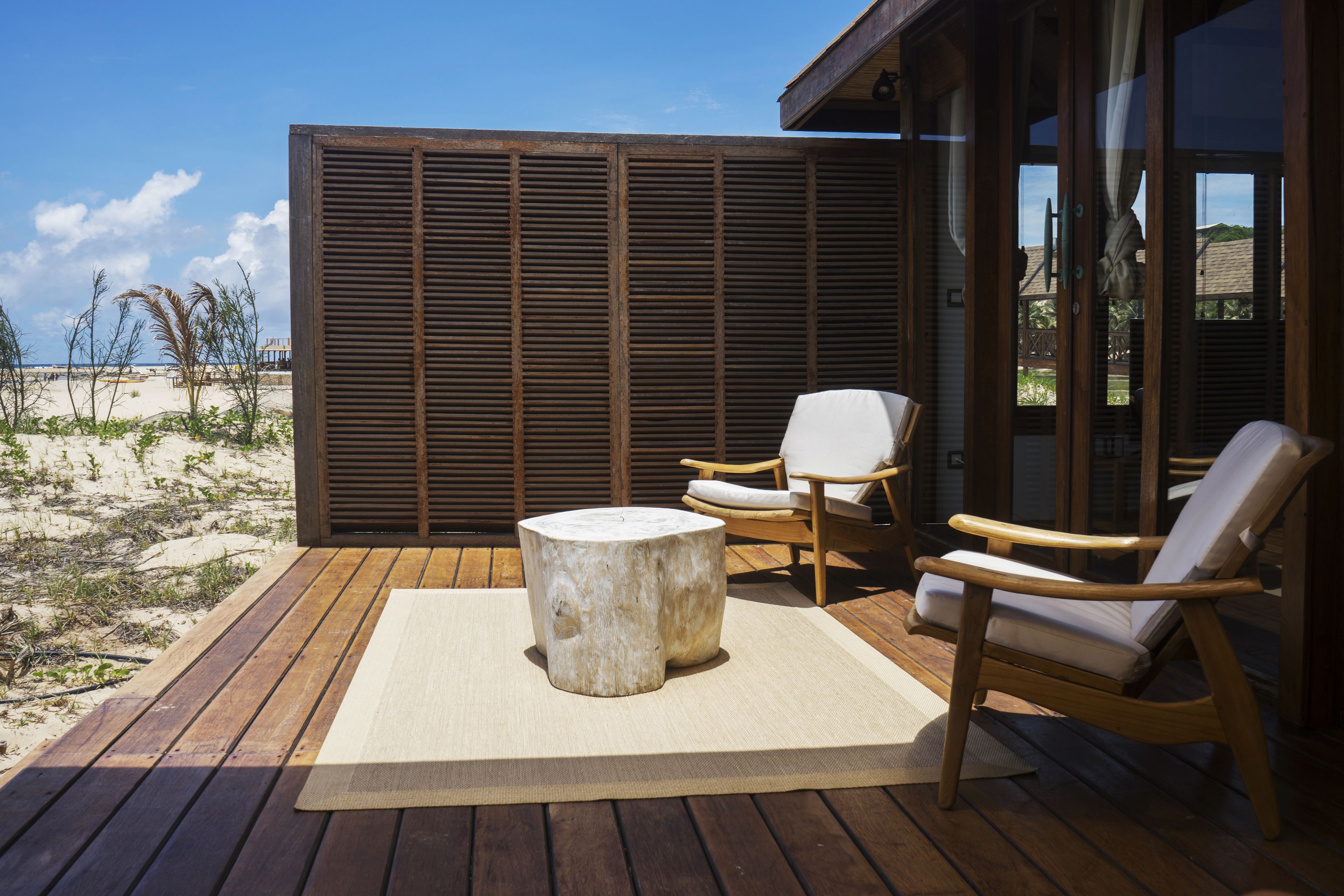 A wooden deck with a round table and two chairs, a large wooden privacy screen, and a view of the beach.