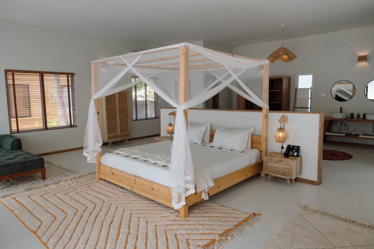 A bedroom with a four-poster bed, a bedside table with a lamp, and a large window with blinds.