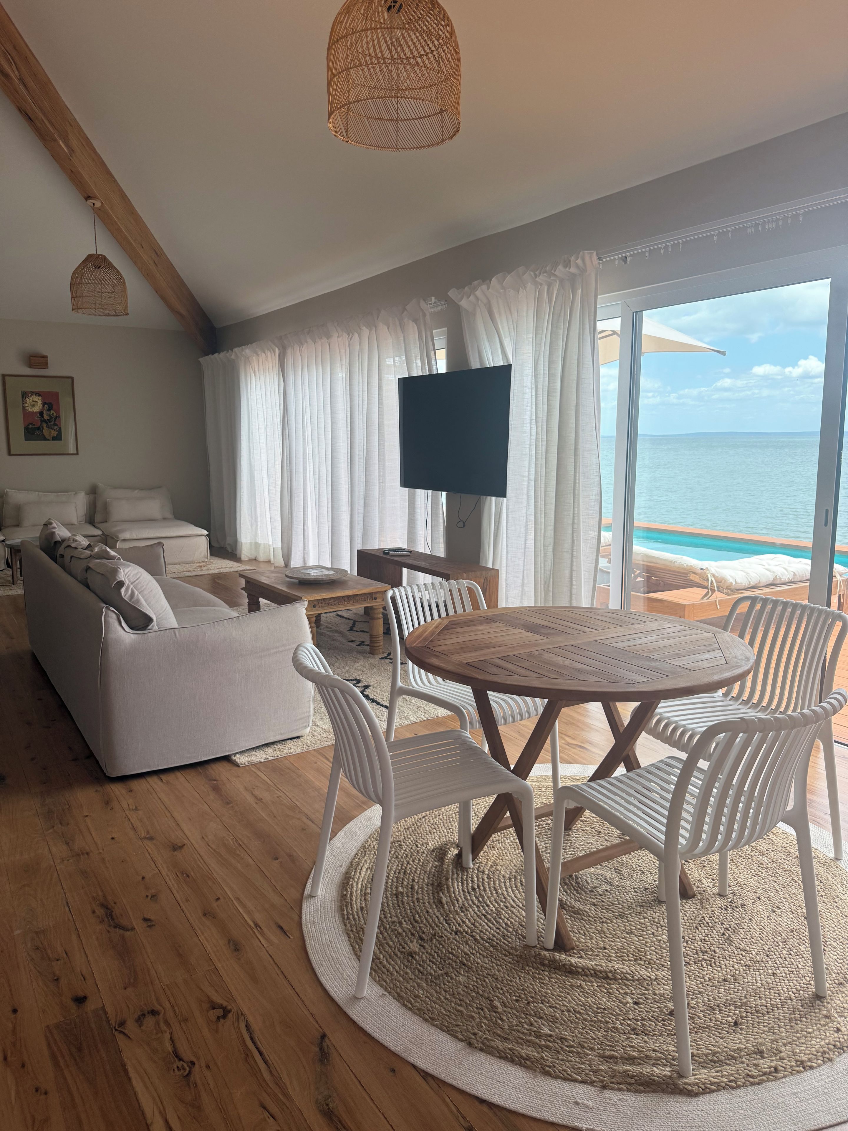 A modern living room with a wooden floor, white furniture, a round wooden table with chairs, a flat-screen TV, and a view of the ocean through large windows.