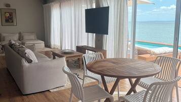 A modern living room with a wooden floor, white furniture, a round wooden table with chairs, a flat-screen TV, and a view of the ocean through large windows.