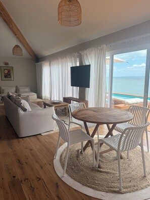 A modern living room with a wooden floor, white furniture, a round wooden table with chairs, a flat-screen TV, and a view of the ocean through large windows. - Sentidos Beach Retreat - Design Hotels (Inhambane)