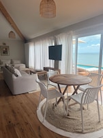 Una sala de estar moderna con piso de madera, mobiliario blanco, una mesa redonda de madera con sillas, un televisor de pantalla plana y vista al océano a través de amplios ventanales.