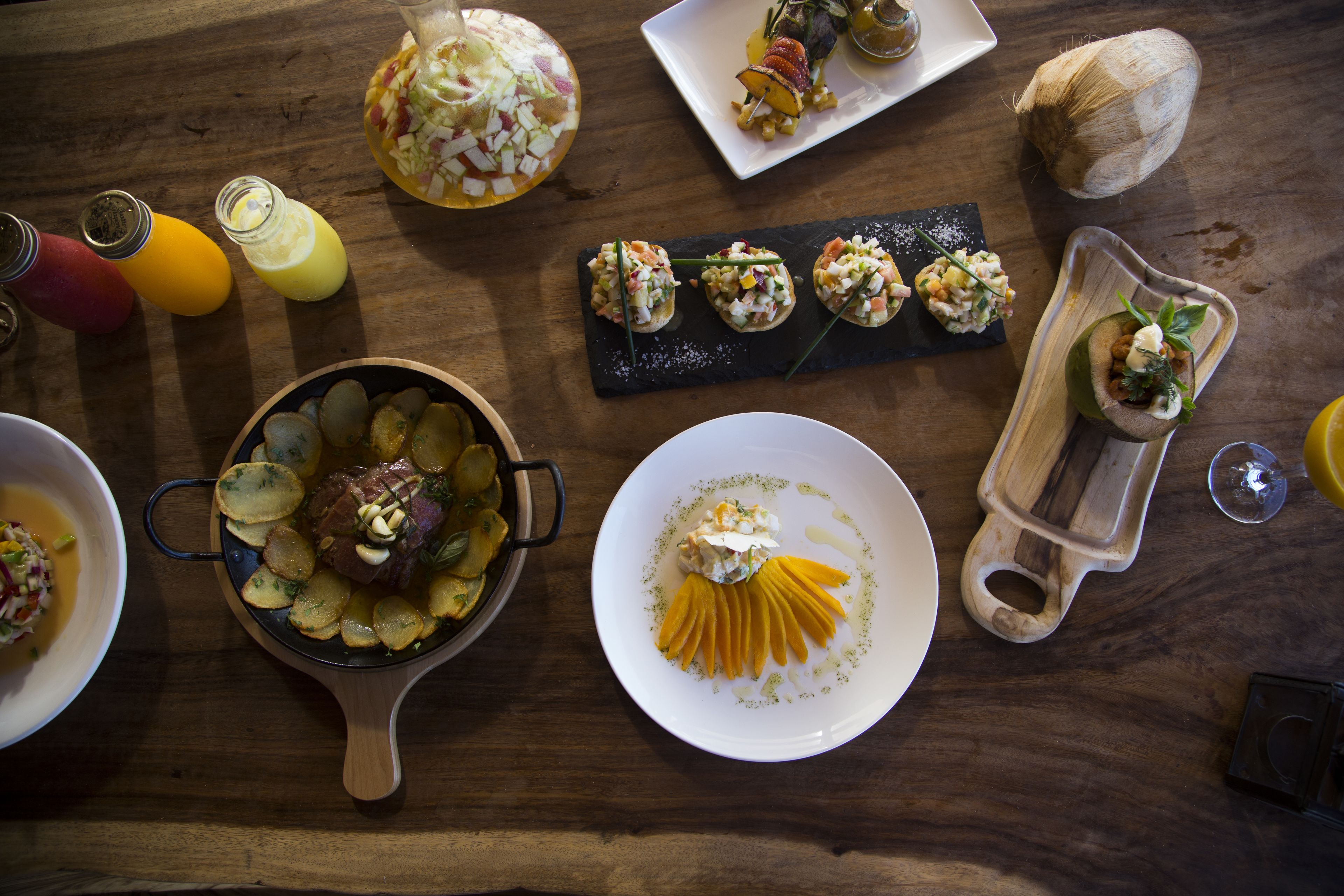 A variety of dishes including a steak with potatoes, a mango salad, and several appetizers on a wooden table.