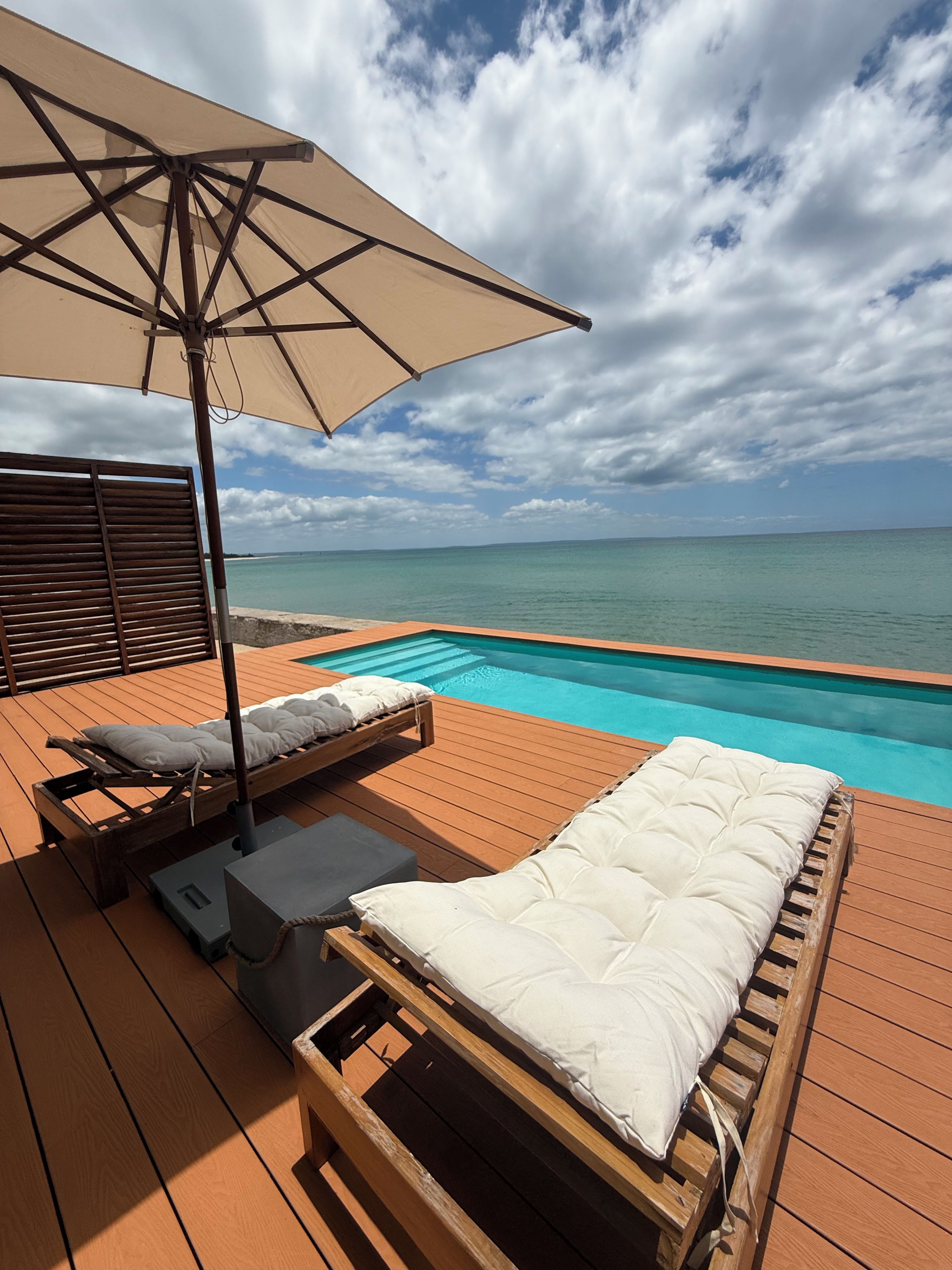 A wooden deck with two sun loungers, one with a beige cushion and the other with a white cushion, under a large umbrella.