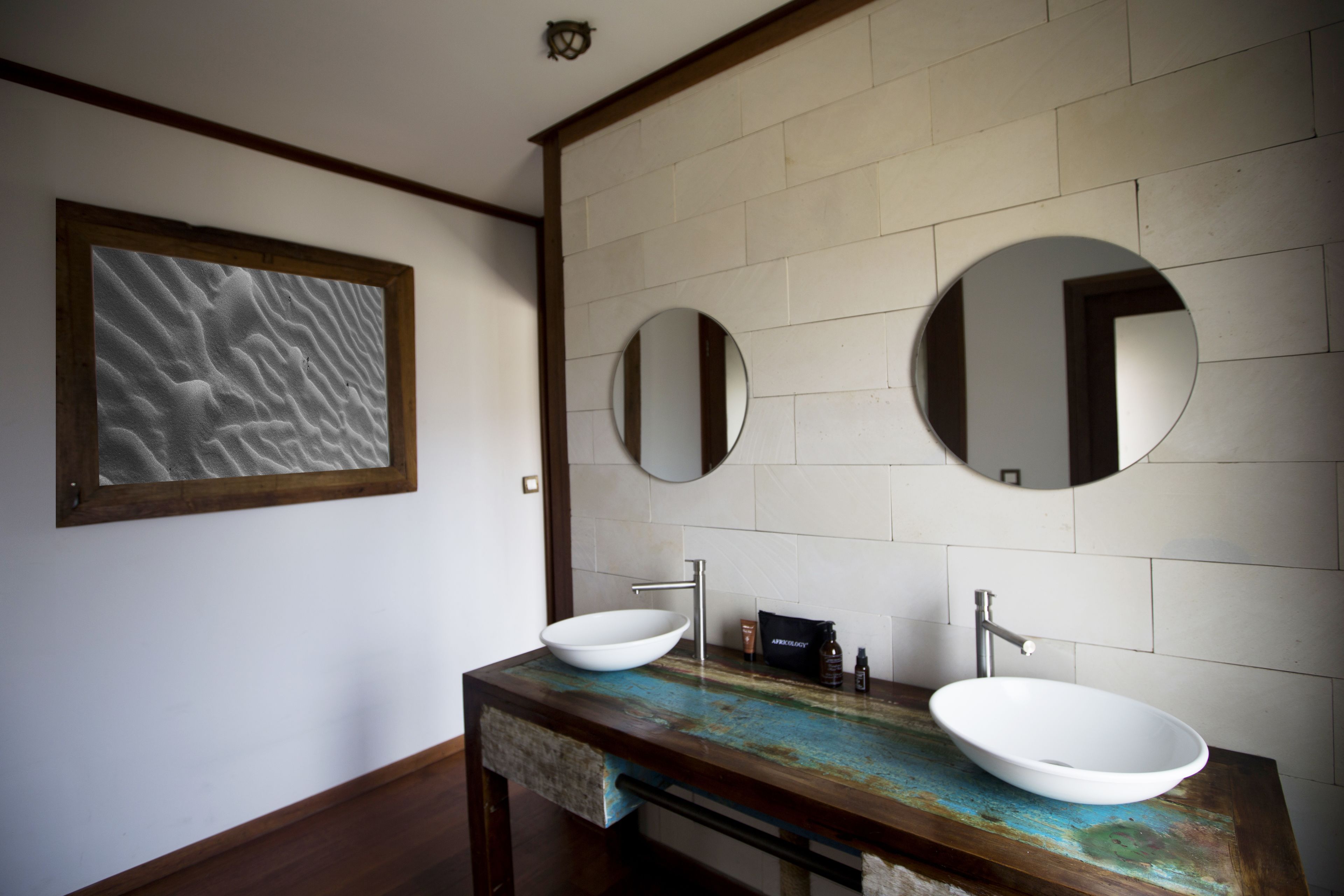 A bathroom with a wooden vanity, two round mirrors, a white sink, and a framed abstract artwork on the wall.