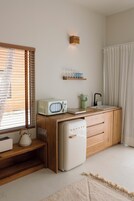 Una cocina compacta con muebles de madera, un microondas blanco, un refrigerador pequeño y una ventana con persianas.