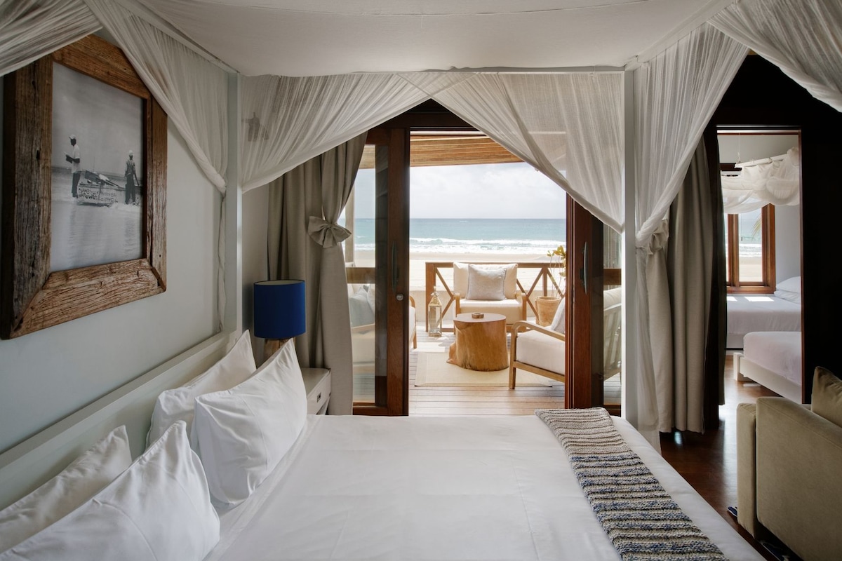 A bedroom with a large bed, a view of the beach, and a balcony with seating.