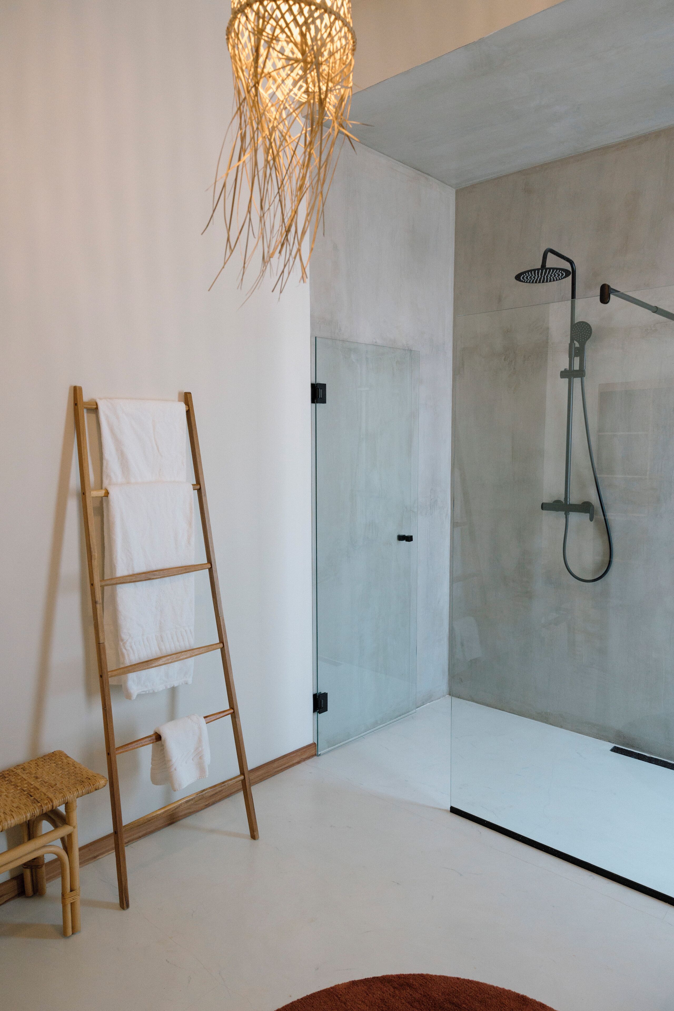 A modern bathroom with a concrete shower wall, a wooden ladder with towels, and a woven light fixture.