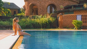 Una mujer sentada junto a una piscina, con un edificio de piedra al fondo.