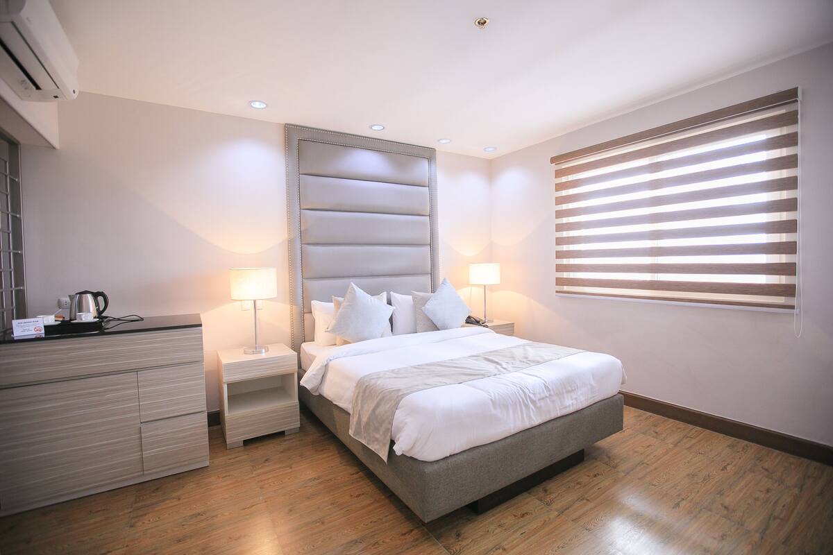A modern bedroom with a bed, nightstands, a dresser, and a window with blinds.