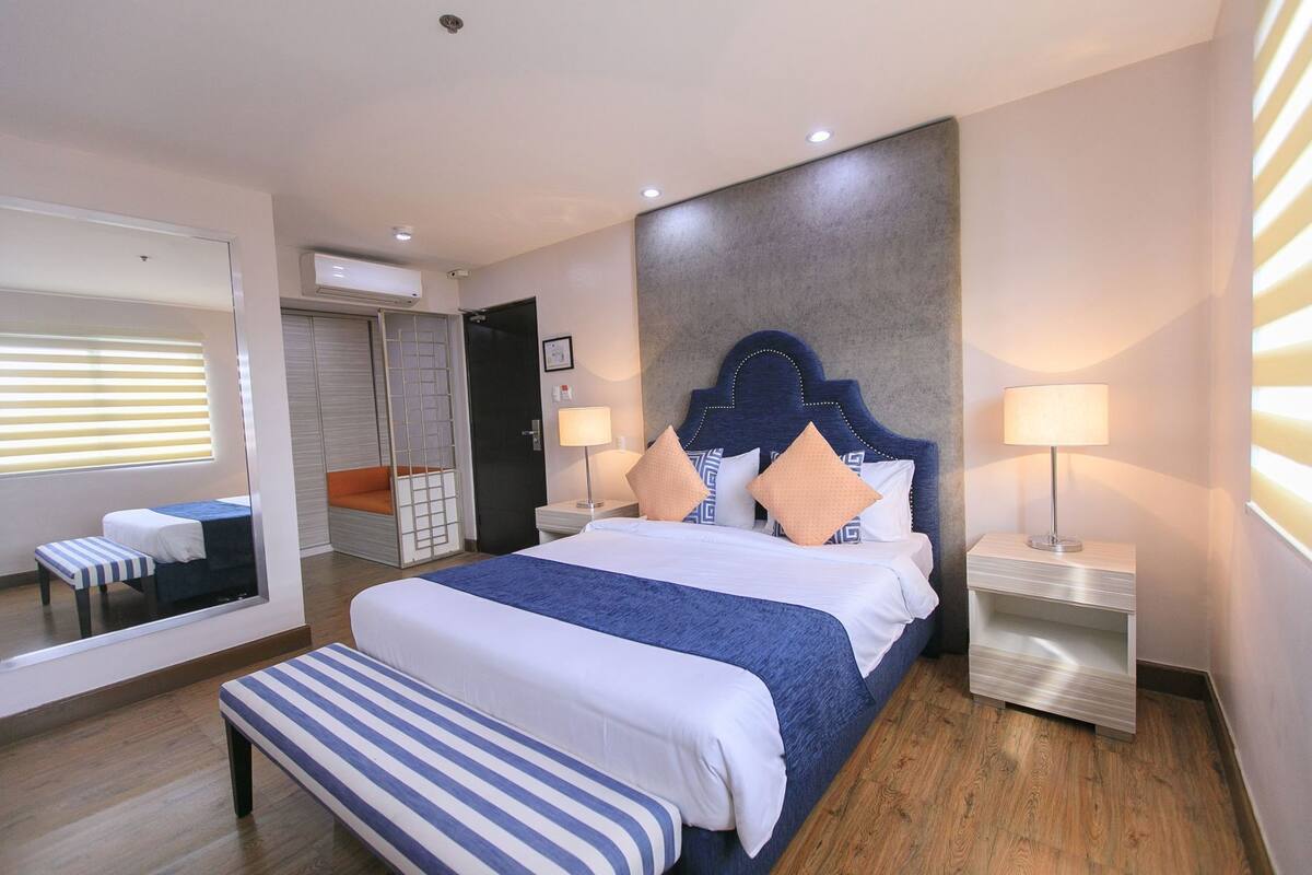 A modern hotel room with a large bed, bedside tables, a bench, a mirror, and a view of the outside.