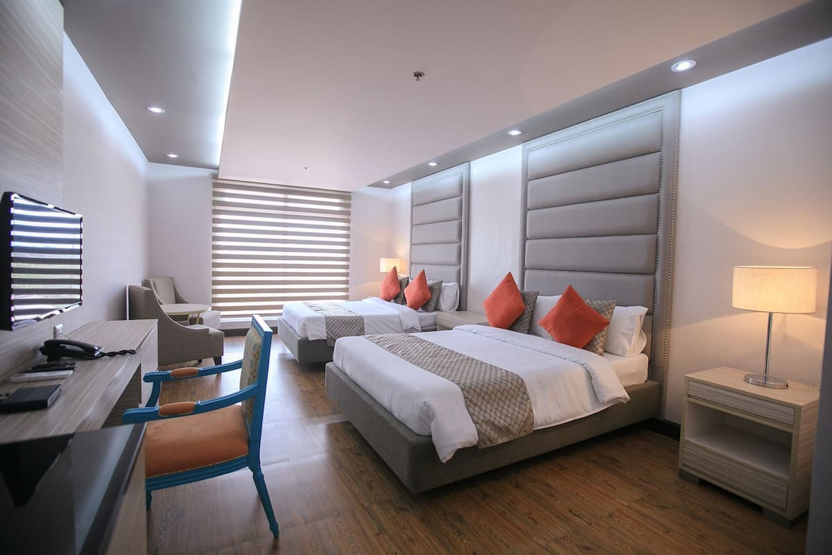 A modern hotel room with a desk, two beds, a chair, and a bedside table.