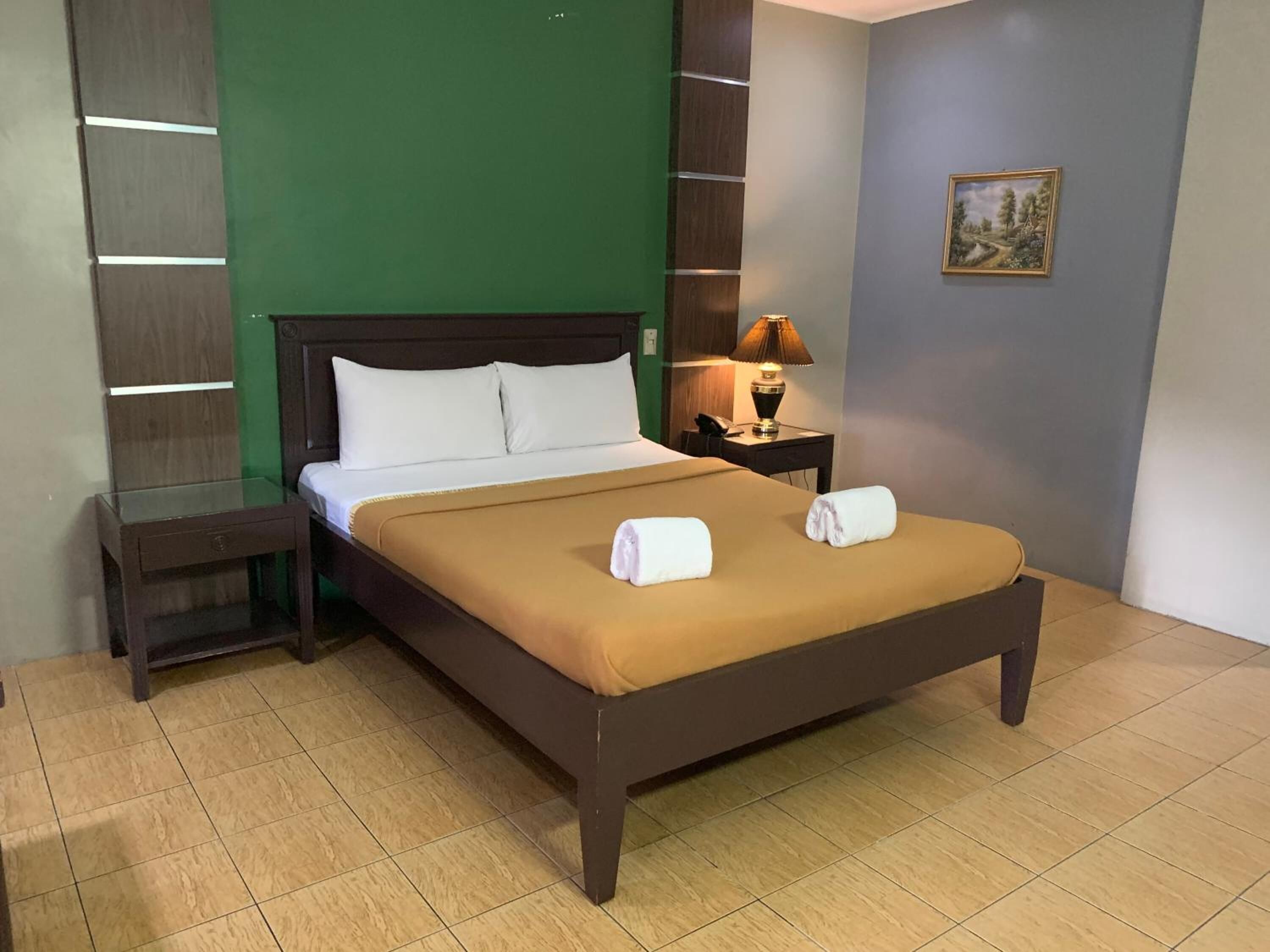 A neatly made bed with white linens and two towels, a bedside table with a lamp, and a painting on the wall.