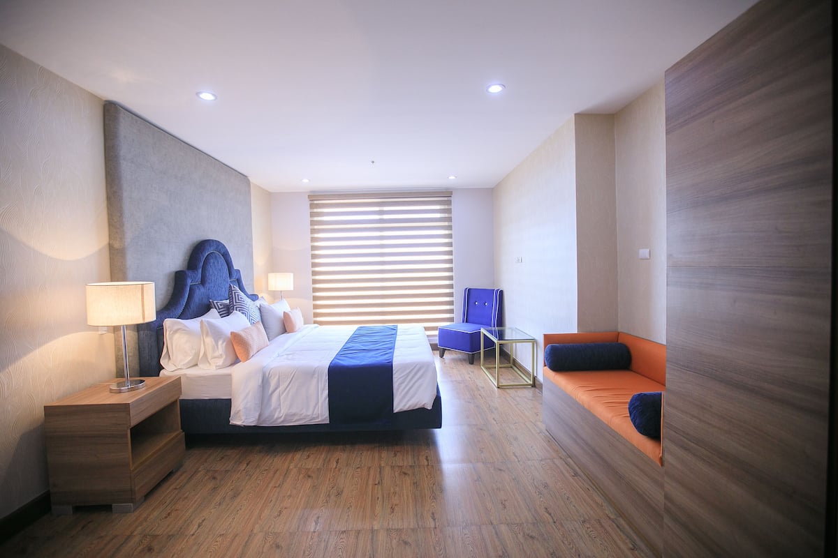 A modern hotel room with a large bed, a bedside table with a lamp, a seating area with a blue chair, and a large window with blinds.