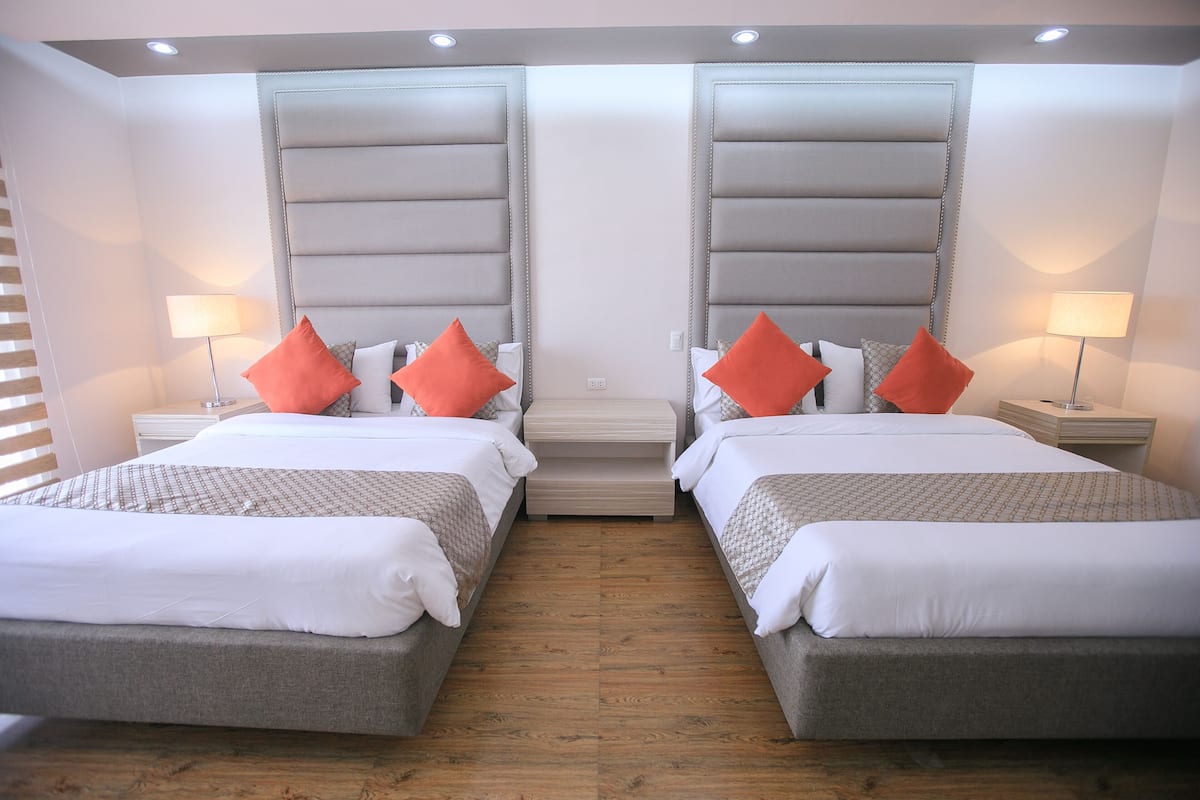 A hotel room with two beds, each with a grey headboard, white bedding, and orange pillows. There are bedside tables with lamps, and the room has wooden flooring.