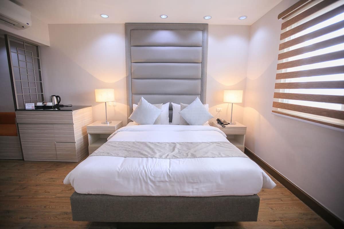 A neatly made bed with a headboard, two bedside tables with lamps, a small dresser, and a window with blinds.