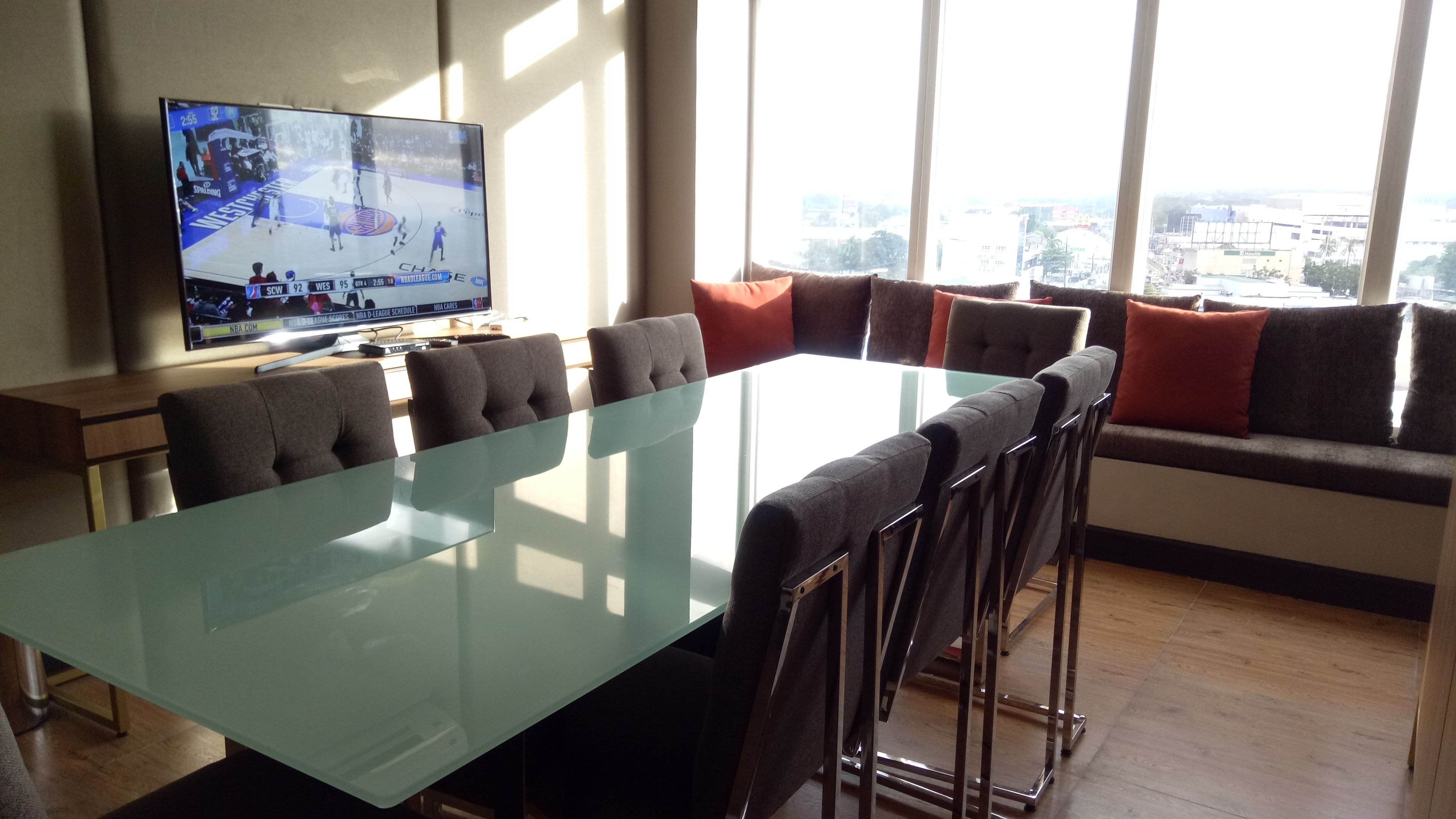 Una sala de conferencias moderna con una mesa de vidrio, asientos cómodos, un televisor grande mostrando un evento deportivo y vistas de la ciudad a través de amplias ventanas.