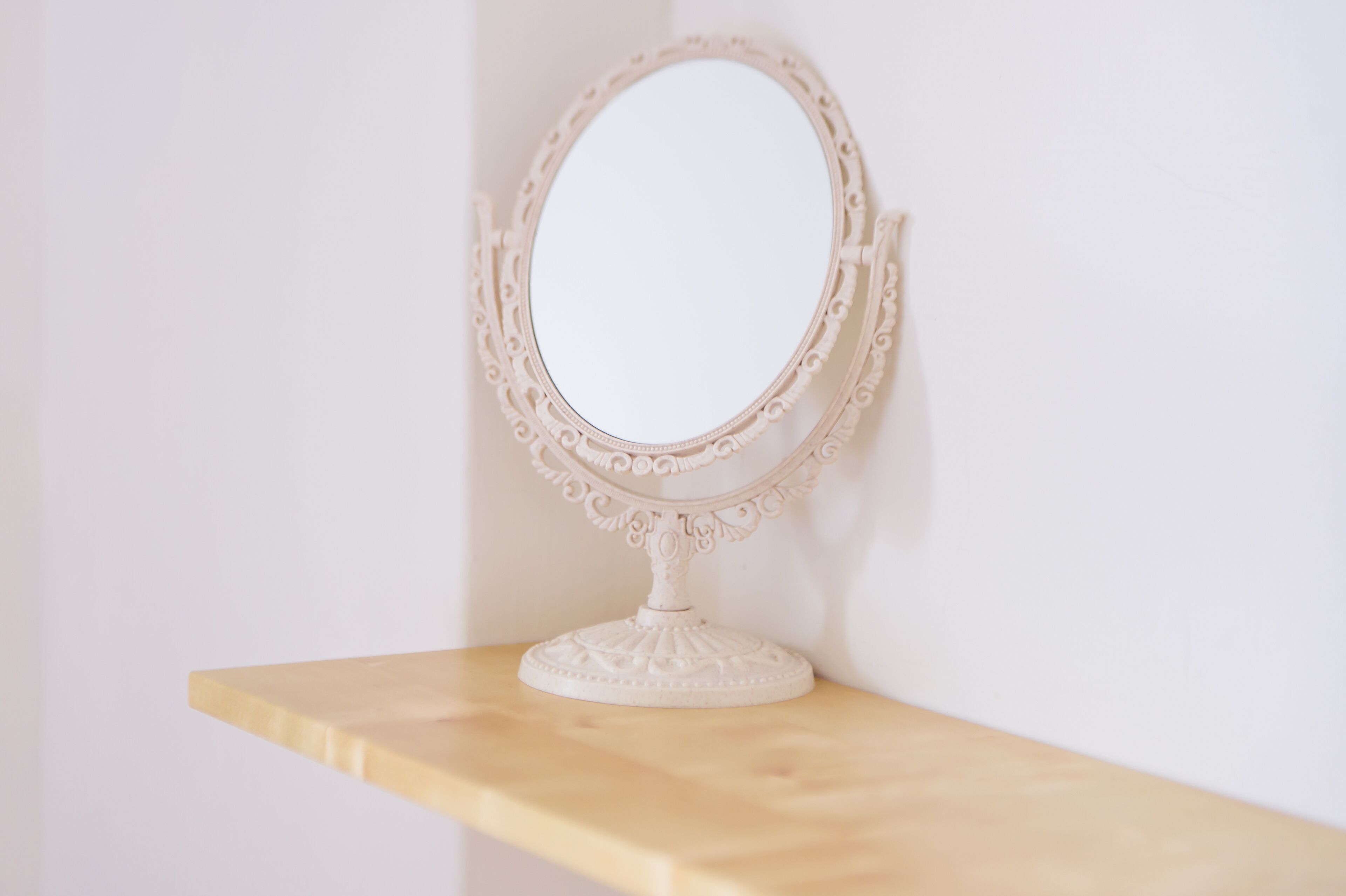 A white ornate mirror with a decorative frame on a wooden shelf against a plain wall.