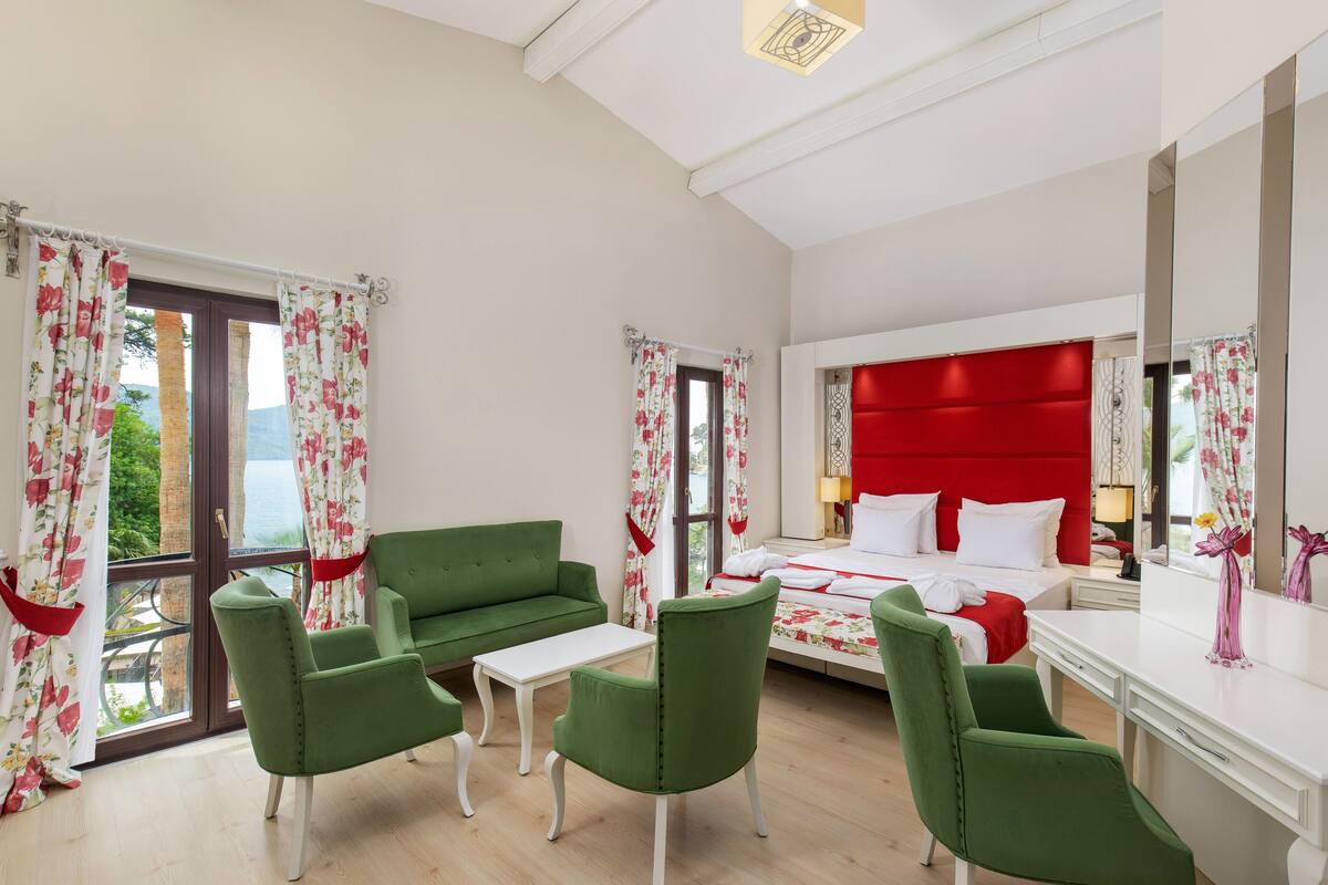 A hotel room with a bed, green armchairs, a white desk, and a large window with curtains.