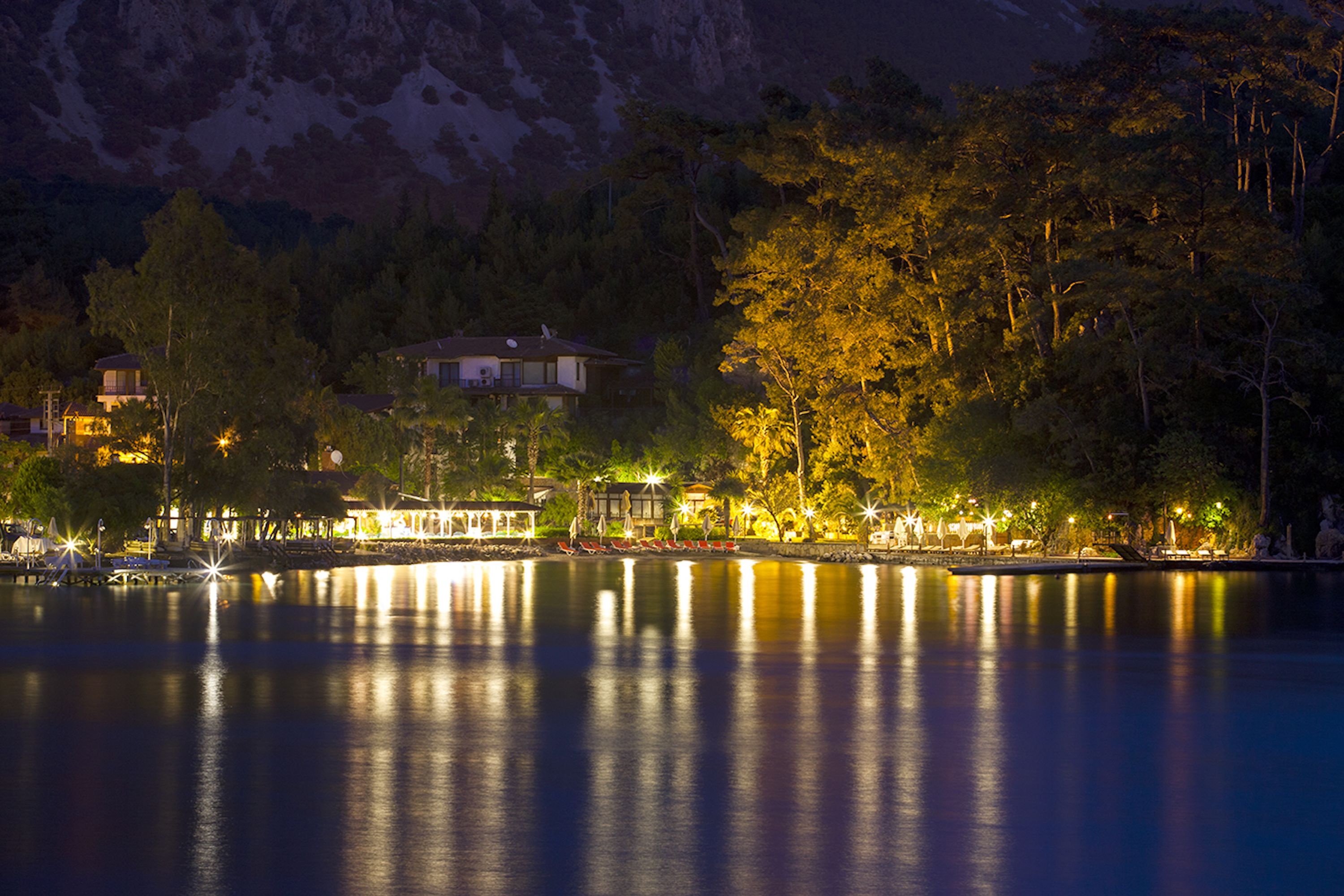 A lakeside view at night with illuminated buildings and trees reflecting on the water.