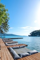 Una terraza de madera con sillones y cojines junto a un lago tranquilo, un muelle que se adentra en el agua y una montaña al fondo.