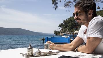 A table with a tray of silverware and a cup, a smartphone on the table, and a view of the sea and hills in the background.