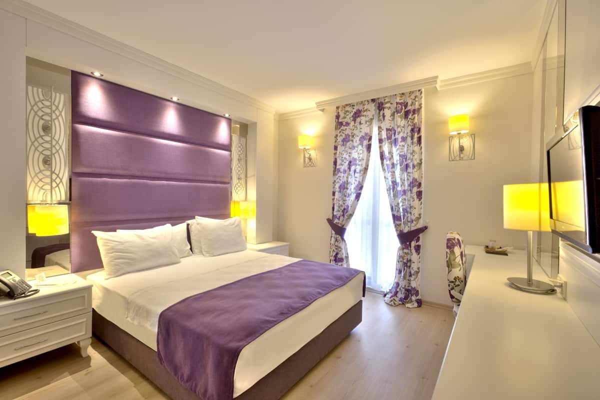 A modern hotel room with a large bed, a bedside table with a lamp, a flat-screen TV, and a decorative wall piece.